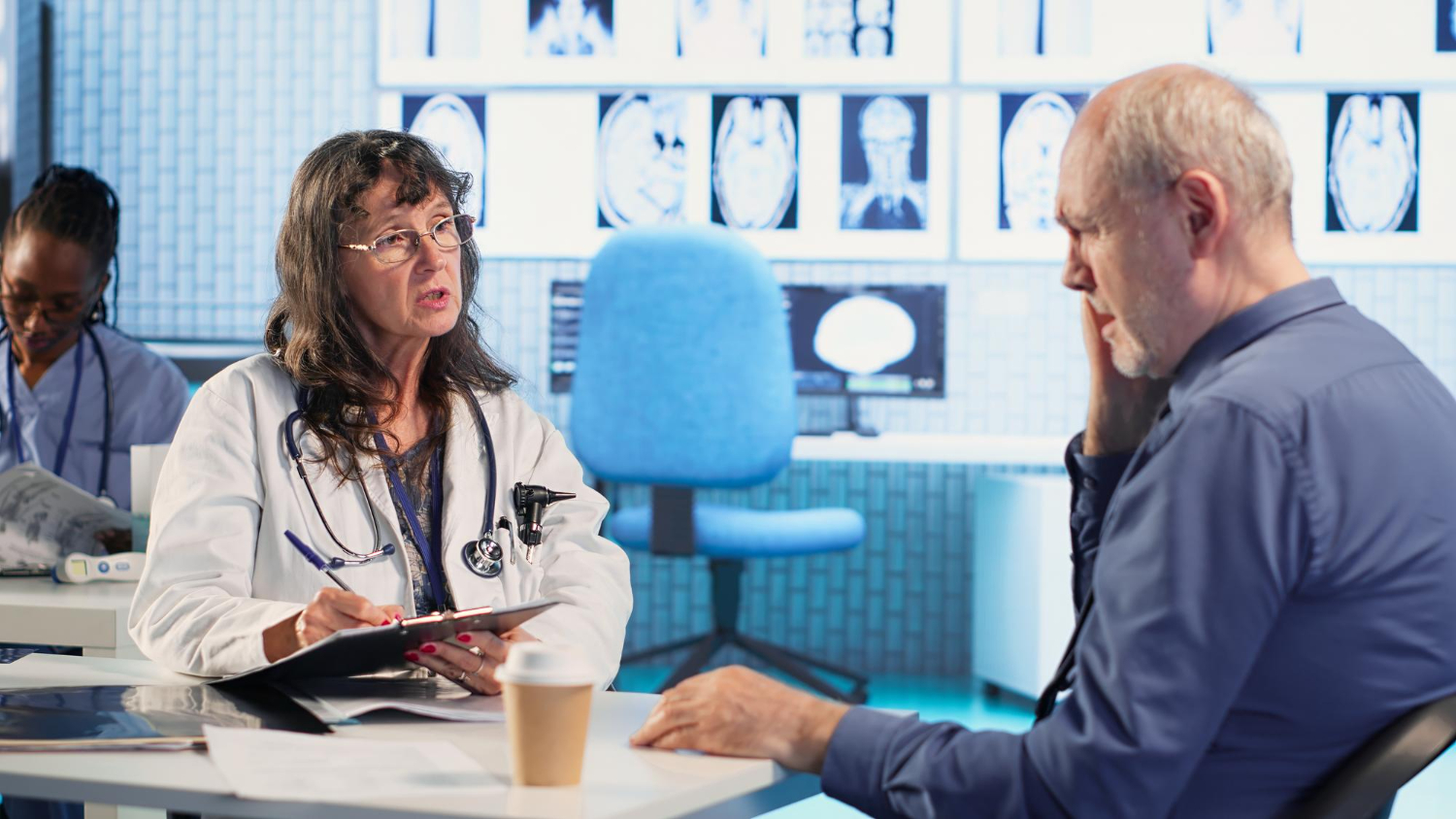 A doctor explains diagnostic imaging results to an older male patient in a medical consultation room, with brain scan images displayed on the wall in the background.