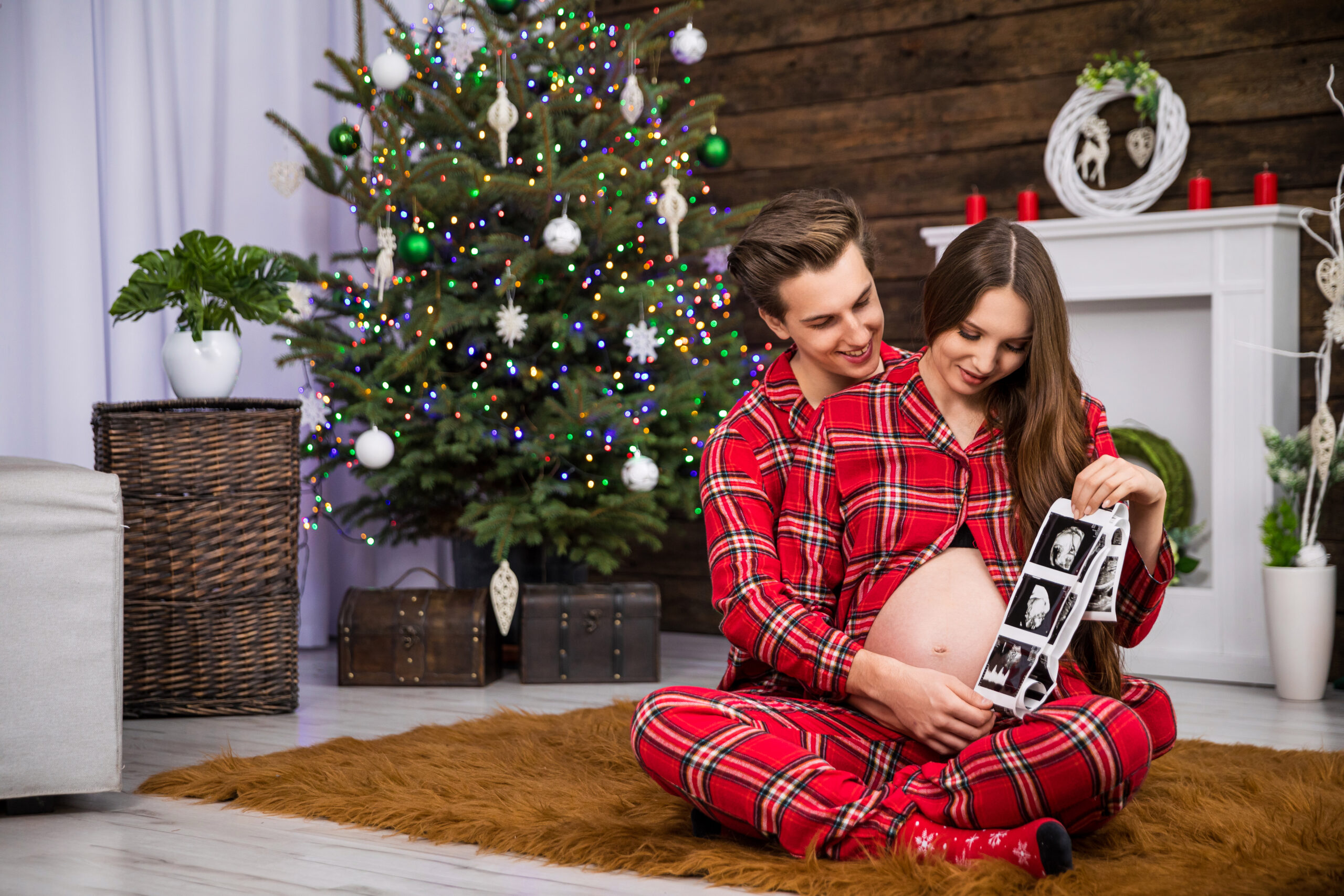 A couple in matching red plaid pajamas sitting by a Christmas tree, joyfully looking at an ultrasound image of their baby together, celebrating the holiday season.