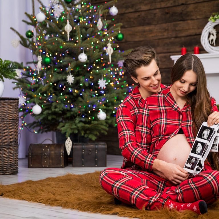 A couple in matching red plaid pajamas sitting by a Christmas tree, joyfully looking at an ultrasound image of their baby together, celebrating the holiday season.